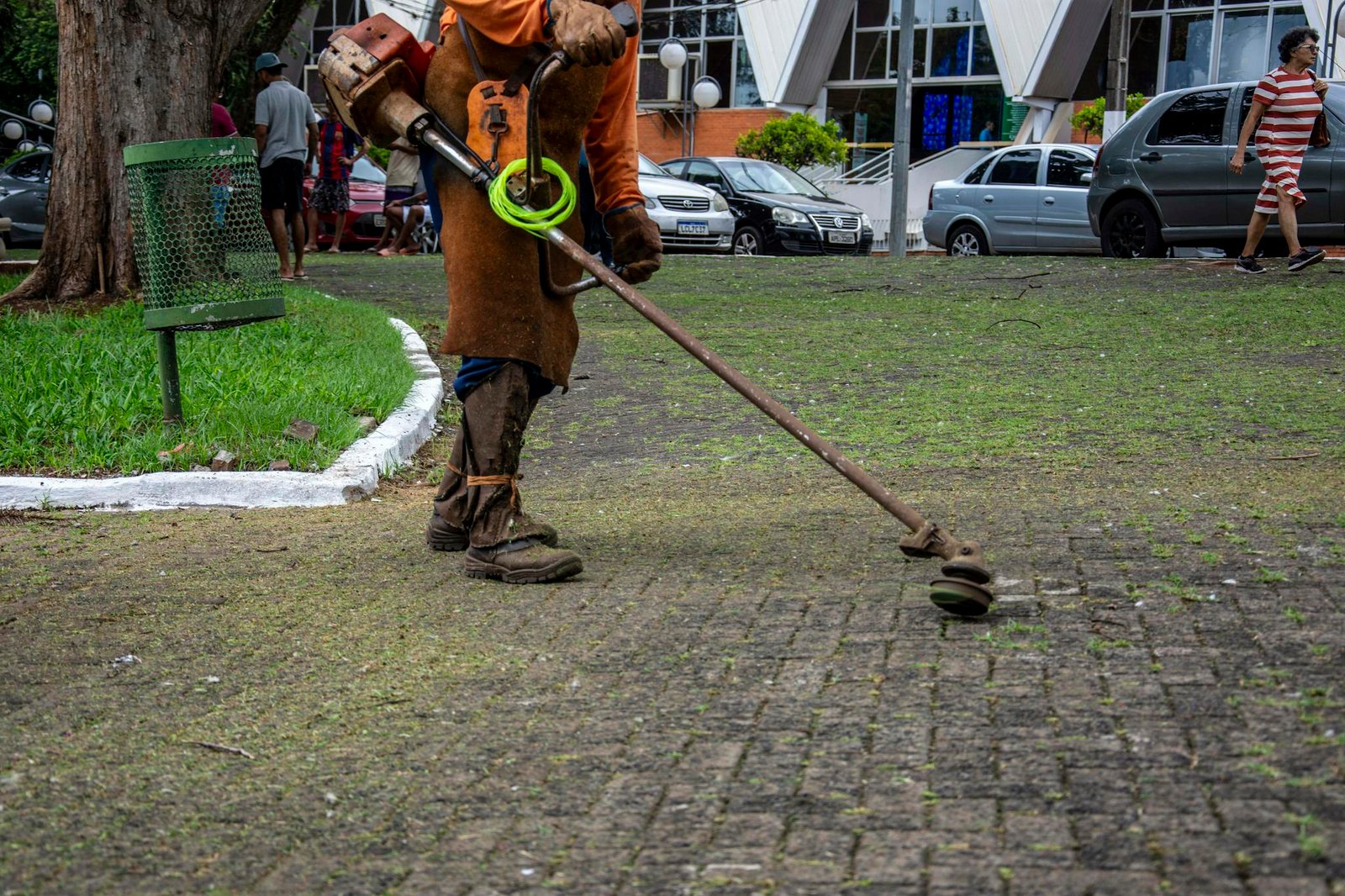 A worker using a trimmer maintains a park in Londrina, Brazil, surrounded by pedestrians and parked cars.